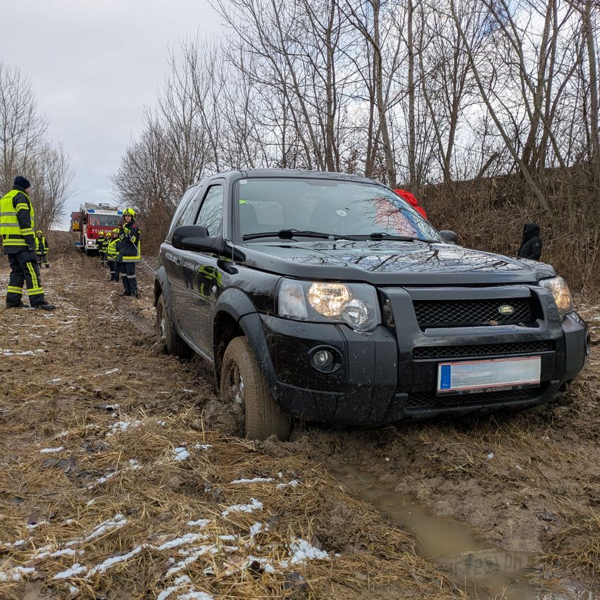 Ein schwarzer SUV ist im schlammigen Gelände steckengeblieben, mit Feuerwehrleuten in der Nähe. Ein Feuerwehrmann steht links, der andere rechts, beide in gelben und schwarzen Uniformen. Ein Feuerwehrwagen ist hinter dem SUV geparkt.