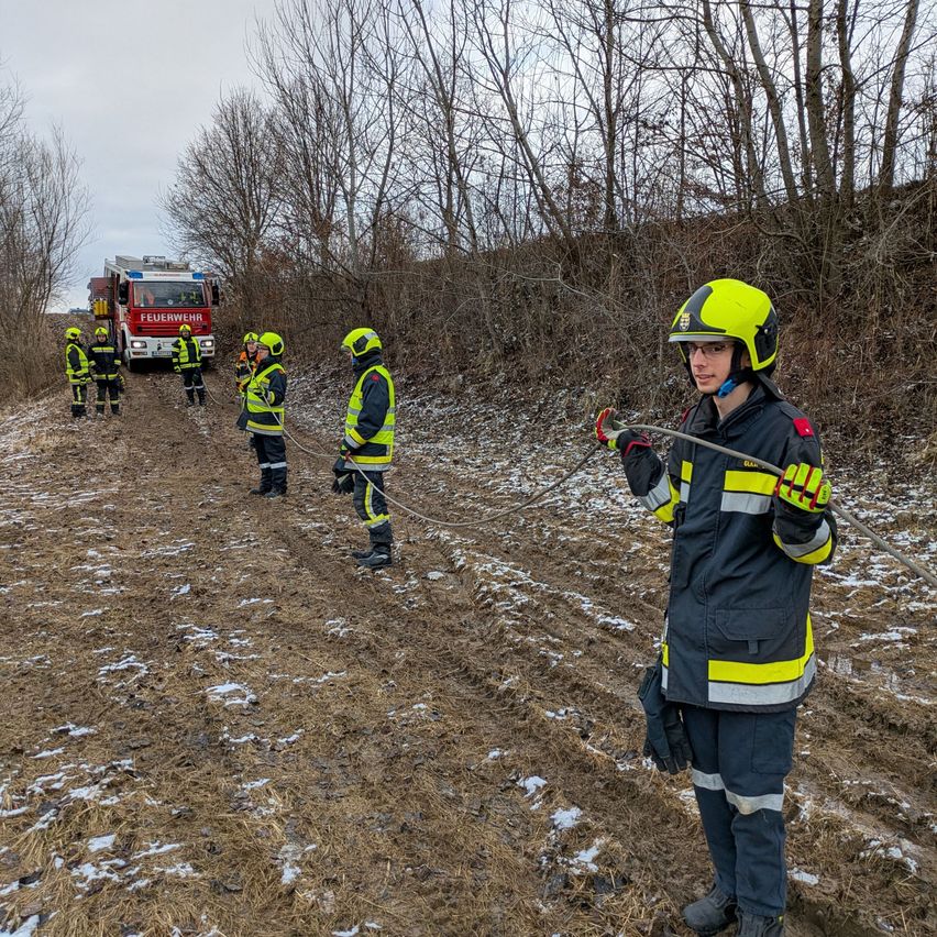 Eine Gruppe von Feuerwehrleuten in gelben und schwarzen Uniformen arbeitet auf einem schlammigen Feld, möglicherweise für eine Trainingsübung. Ein Feuerwehrwagen ist in der Nähe geparkt.