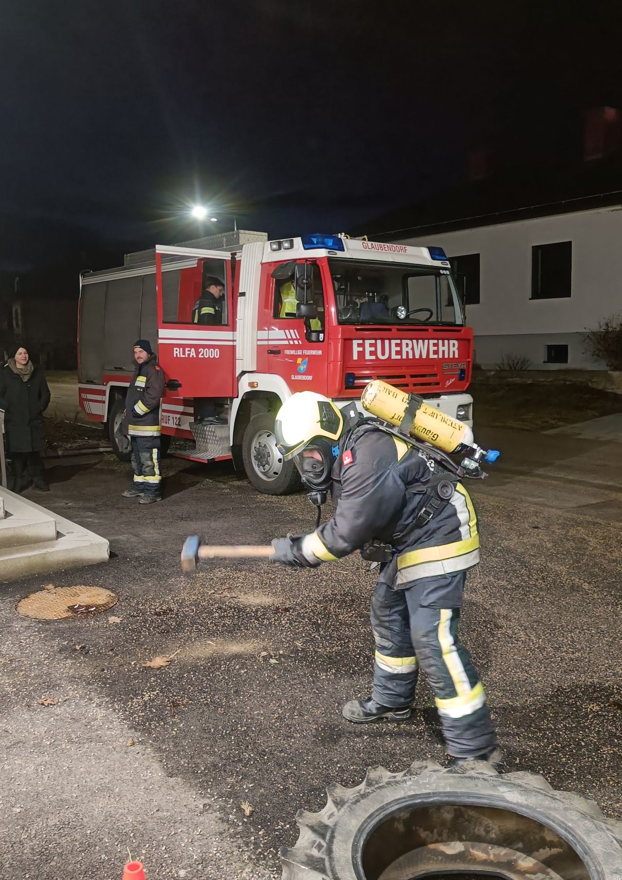 Ein Feuerwehrmann in voller Ausrüstung ist zu sehen, wie er vor einem Feuerwehrauto steht, einen Hammer hält und den Boden bricht. Zwei weitere Feuerwehrleute sind anwesend, einer steht neben dem Auto. Die Szene spielt sich bei Nacht ab.
