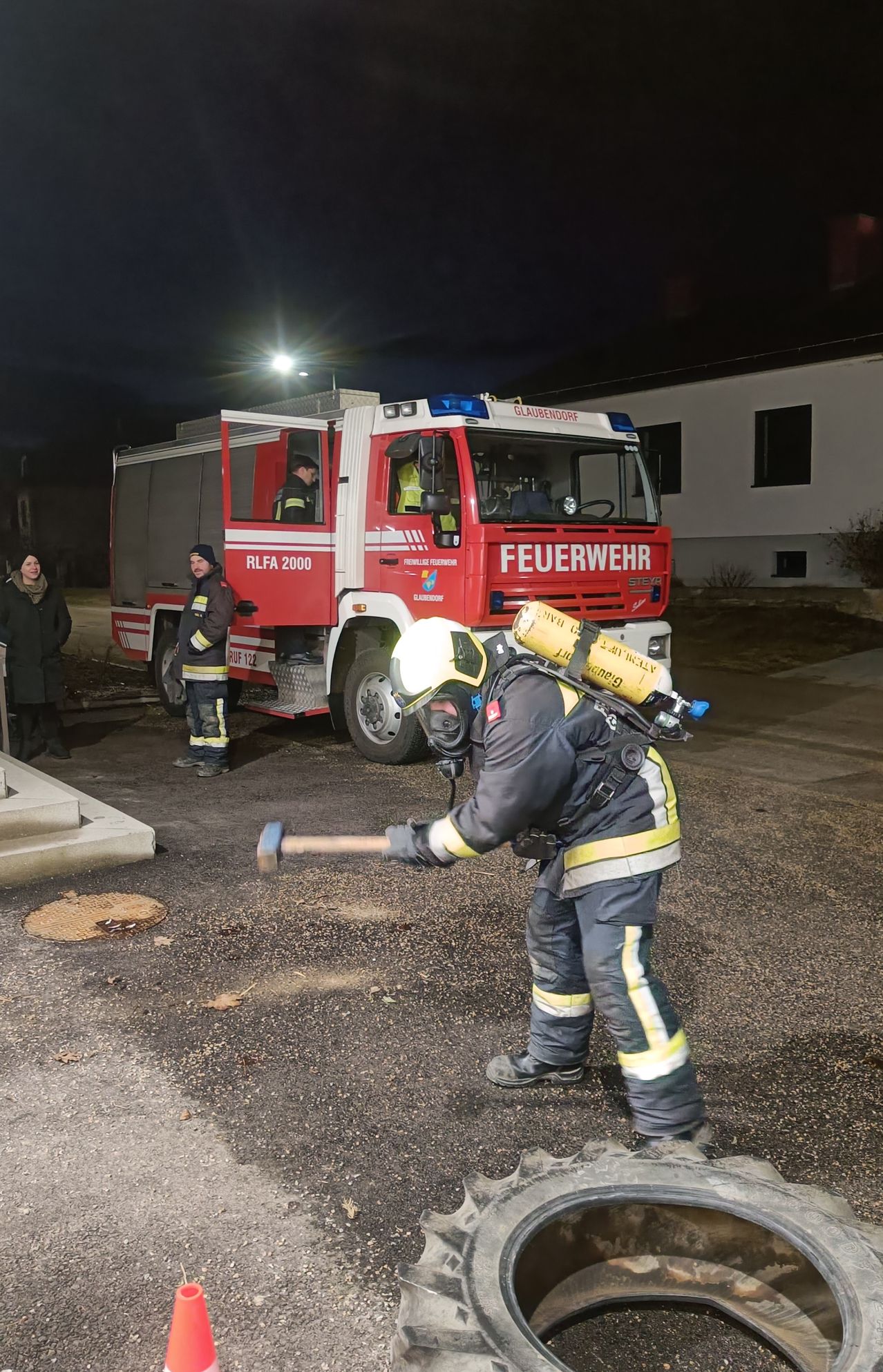 Ein Feuerwehrmann in voller Ausrüstung ist zu sehen, wie er vor einem Feuerwehrauto steht, einen Hammer hält und den Boden bricht. Zwei weitere Feuerwehrleute sind anwesend, einer steht neben dem Auto. Die Szene spielt sich bei Nacht ab.