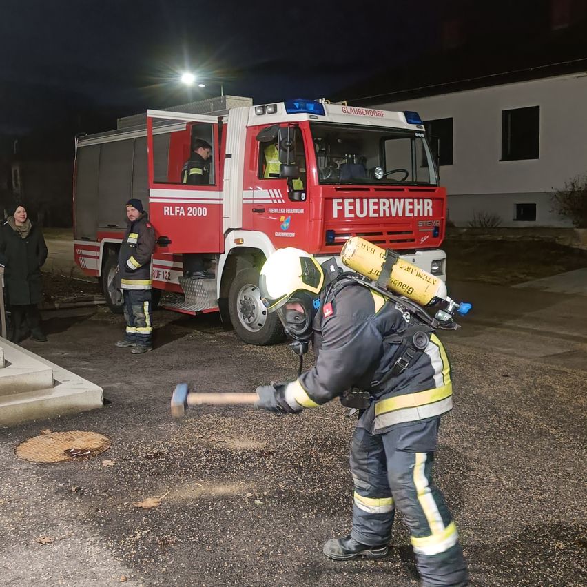 Ein Feuerwehrmann in voller Ausrüstung ist zu sehen, wie er vor einem Feuerwehrauto steht, einen Hammer hält und den Boden bricht. Zwei weitere Feuerwehrleute sind anwesend, einer steht neben dem Auto. Die Szene spielt sich bei Nacht ab.