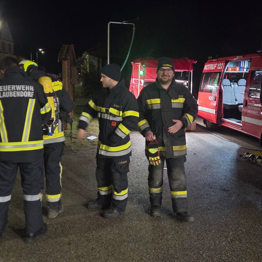 Drei Feuerwehrleute stehen bei Nacht draußen mit einem Feuerwehrwagen in der Nähe. Ein Feuerwehrmann hält einen Helm, und sie sind alle in ihren Uniformen.