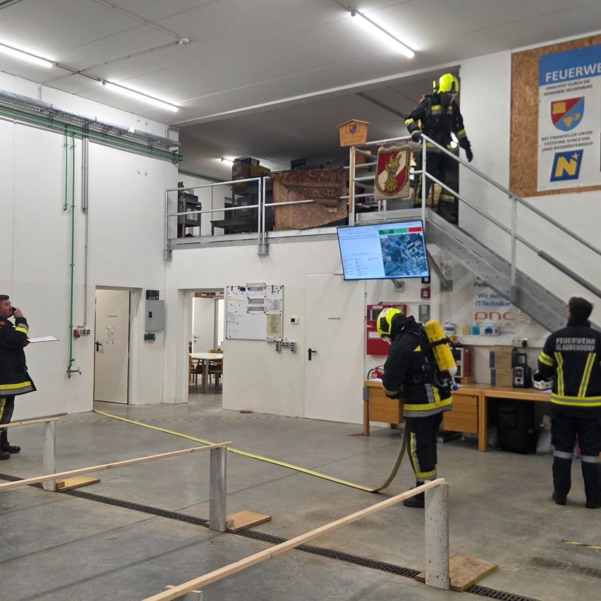 Eine Feuerwache mit zwei Feuerwehrleuten in voller Montur, einer klettert eine Treppe mit einem Schild, ein anderer steht neben einem Monitor, und zwei weitere beobachten. Der Raum hat eine weiße Wand, eine Treppe und einen Tisch mit einem Feuerlöscher.