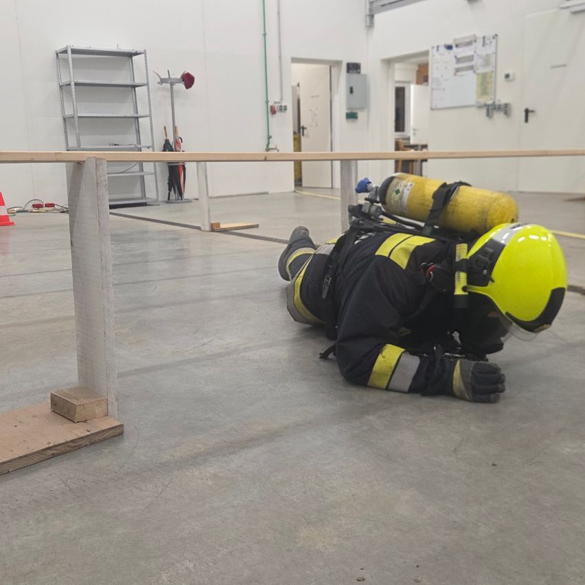 Ein Feuerwehrmann in voller gelber Schutzausrüstung und Helm kriecht auf dem Boden, mit einem gelben Sauerstofftank auf dem Rücken. Der Bereich ist eine Trainingseinrichtung mit weißen Wänden und grauem Boden.