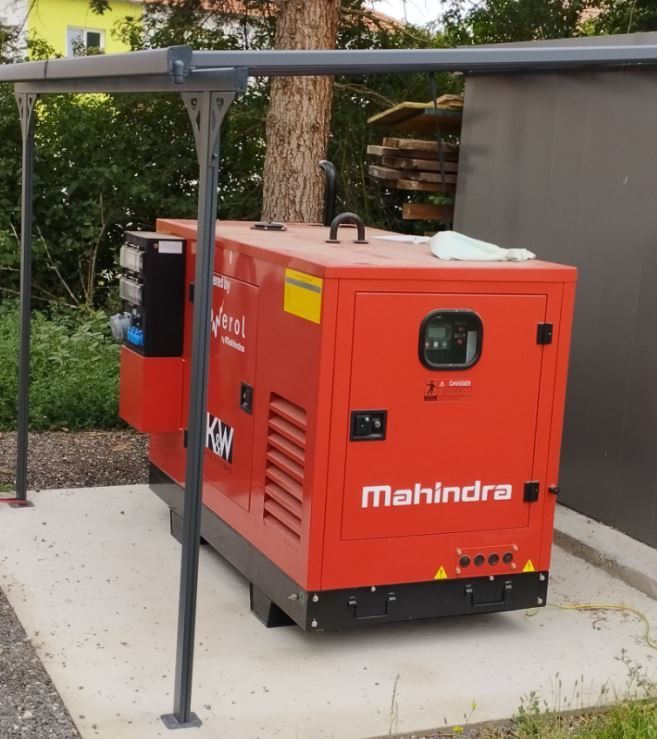 Ein roter Mahindra-Generator steht auf einer Betonfläche, abgedeckt von einem grauen Vordach, mit einem gelb-schwarzen Schild und einem Bedienfeld.