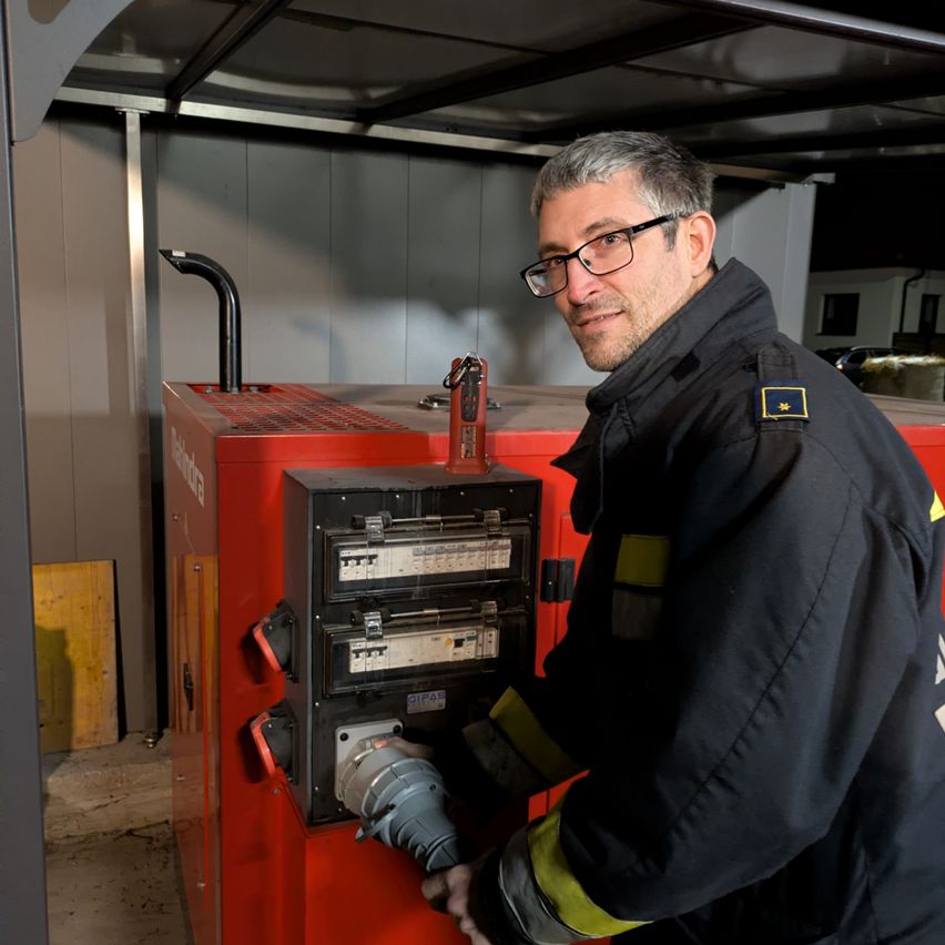 Ein Feuerwehrmann mit Brille und Jacke steht neben einer roten Maschine, an die ein Stromkabel angeschlossen ist.
