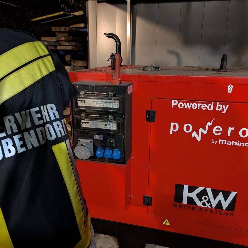 Eine Person in einer Feuerwehruniform steht neben einem roten Generator mit der Aufschrift 'Powered by powero by Mahina'.