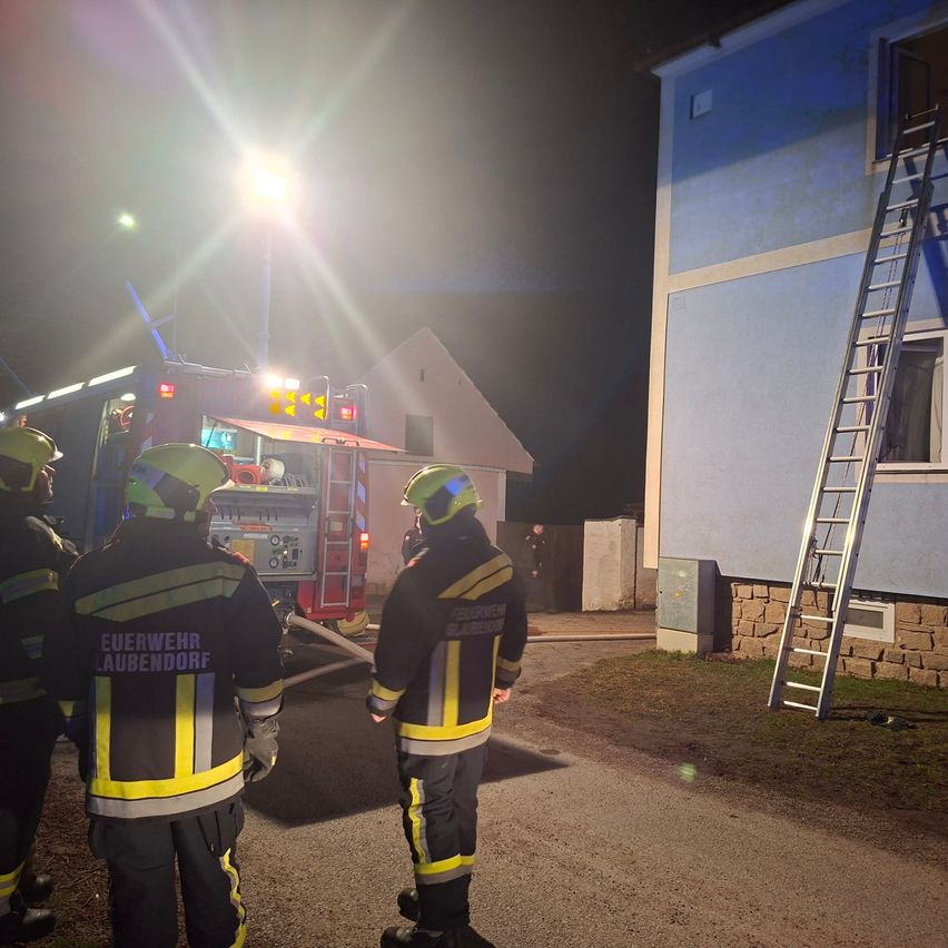 Feuerwehrleute in voller Ausrüstung stehen bei Nacht vor einem Gebäude, mit einem Feuerwehrwagen und einer Leiter in der Nähe. Die Szene wird von den Lichtern des Feuerwehrwagens beleuchtet.