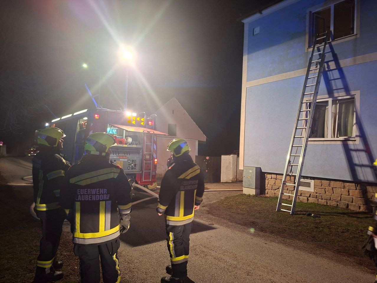 Feuerwehrleute in voller Ausrüstung stehen bei Nacht vor einem Gebäude, mit einem Feuerwehrwagen und einer Leiter in der Nähe. Die Szene wird von den Lichtern des Feuerwehrwagens beleuchtet.