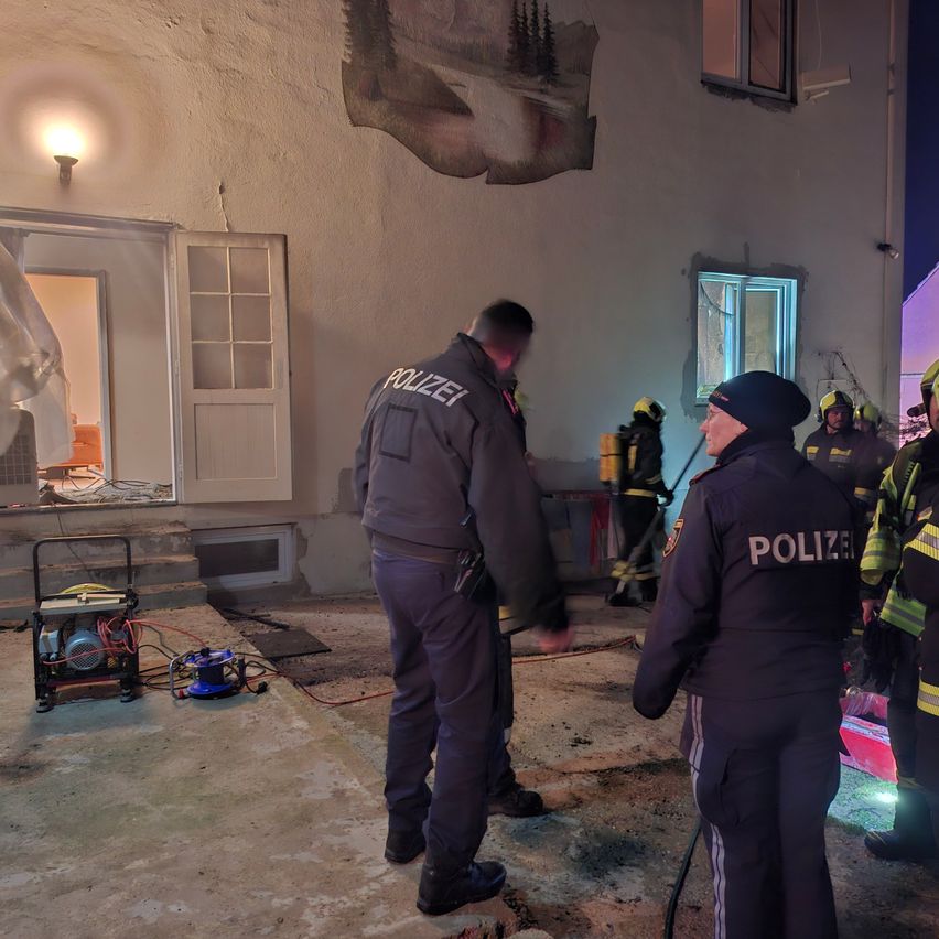 Polizisten und Feuerwehrleute versammeln sich vor einem beschädigten Gebäude. Der vordere Polizist trägt einen Hut und eine Jacke mit dem Wort 'POLIZEI'. Ein Feuerwehrmann benutzt einen Schlauch.