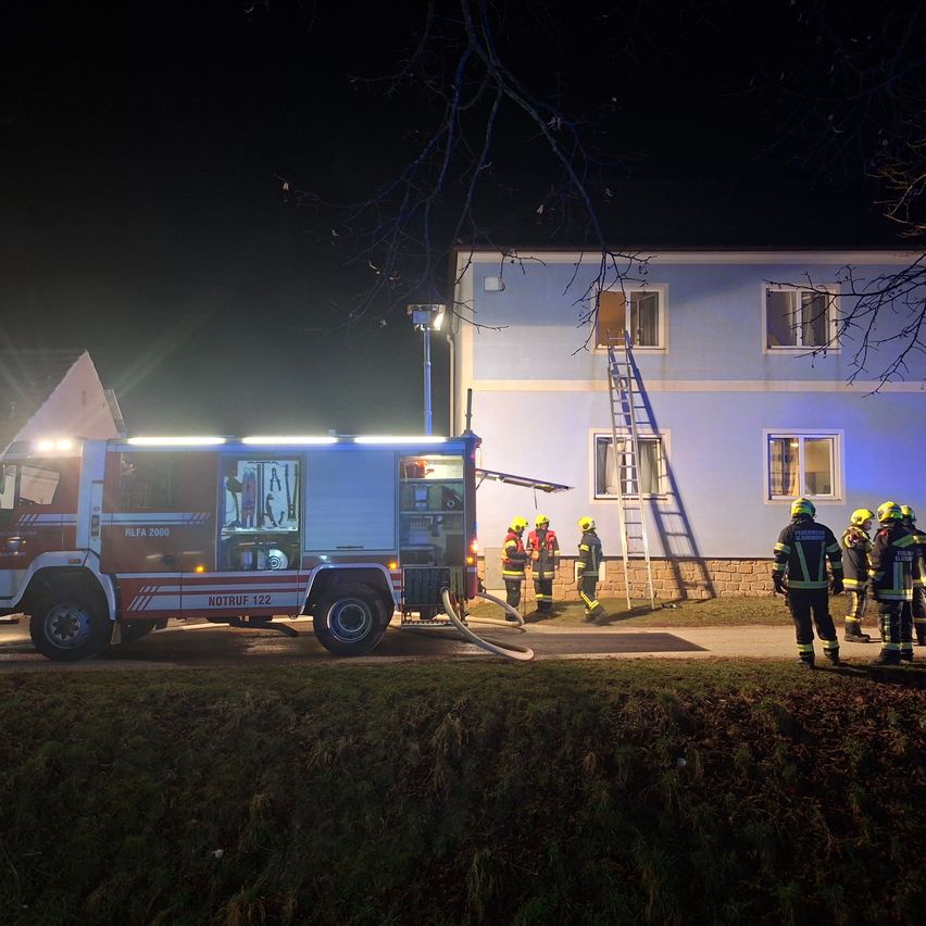 Feuerwehrleute reagieren auf einen Notfall in einem Gebäude. Ein Feuerwehrwagen ist vor dem Gebäude geparkt, und Feuerwehrleute sind in der Nähe versammelt. Eine Leiter ist an das Gebäude gelehnt.