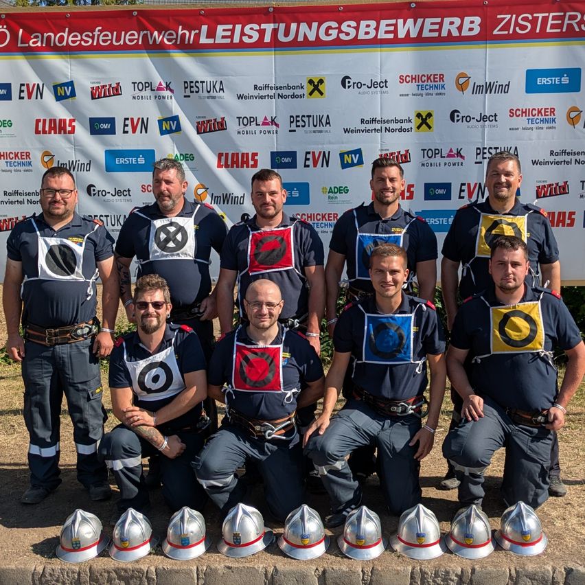 Eine Gruppe von Feuerwehrleuten in Uniform mit Helmen vor ihnen, posiert für ein Foto vor einem Banner mit verschiedenen Logos.