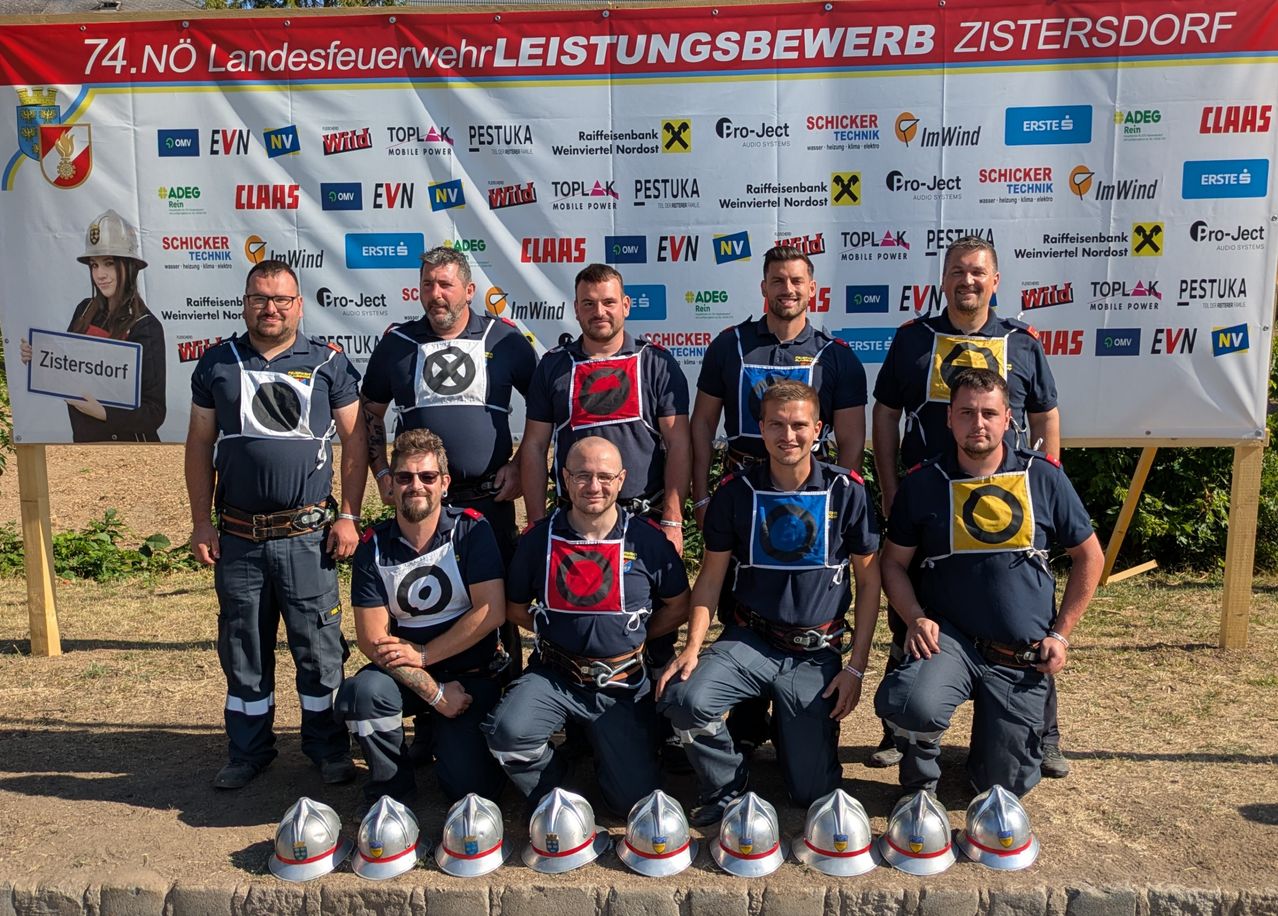 Eine Gruppe von Feuerwehrleuten in Uniform mit Helmen vor ihnen, posiert für ein Foto vor einem Banner mit verschiedenen Logos.