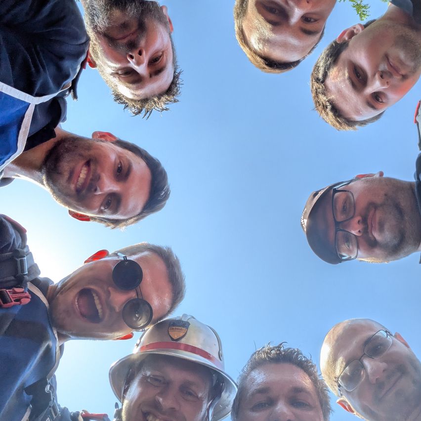Eine Gruppe von Männern lächelt und posiert für ein Foto vor einem blauen Himmel. Einige tragen Brillen und Hüte, einer trägt einen Helm.