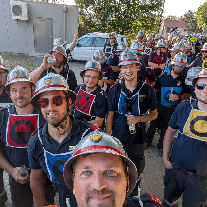 Eine Gruppe von Feuerwehrleuten in Uniform, die Helme tragen, steht zusammen draußen. Einige trinken aus Flaschen und scheinen für ein Foto zu posieren.