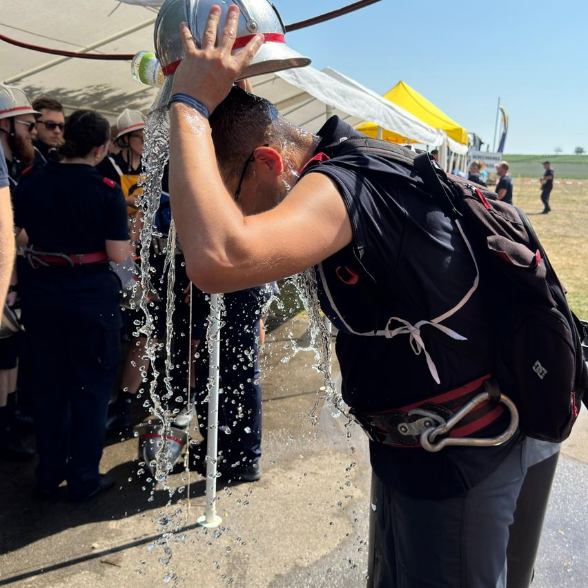 Ein Feuerwehrmann lässt sich von einer anderen Person den Helm mit Wasser füllen, an einem sonnigen Außenort.