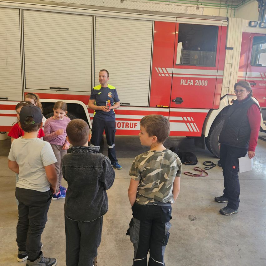 Eine Gruppe von Kindern hört einem Feuerwehrmann vor einem Feuerwehrwagen in einer Garage zu.