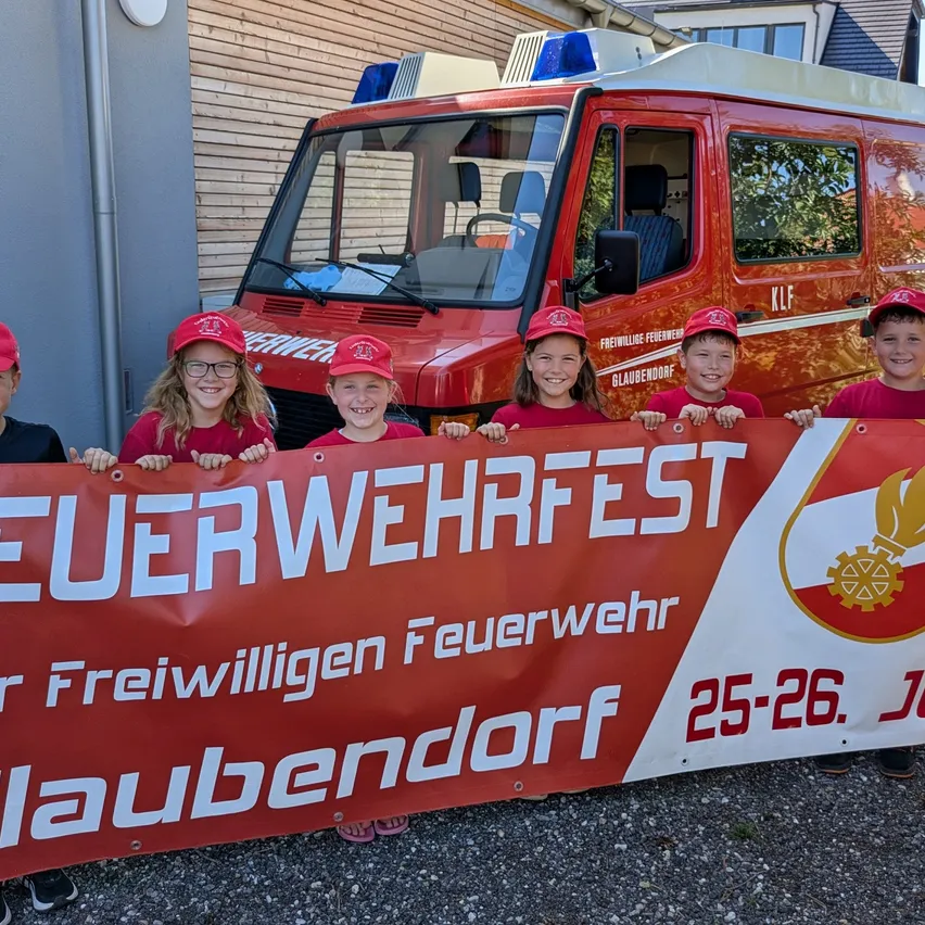 Eine Gruppe von Kindern in roten Mützen und T-Shirts steht hinter einem Banner, das 'Feuerwehrfest' vor einem roten Feuerwehrwagen besagt. Das Banner wird von den Kindern hochgehalten, und sie lächeln für die Kamera.