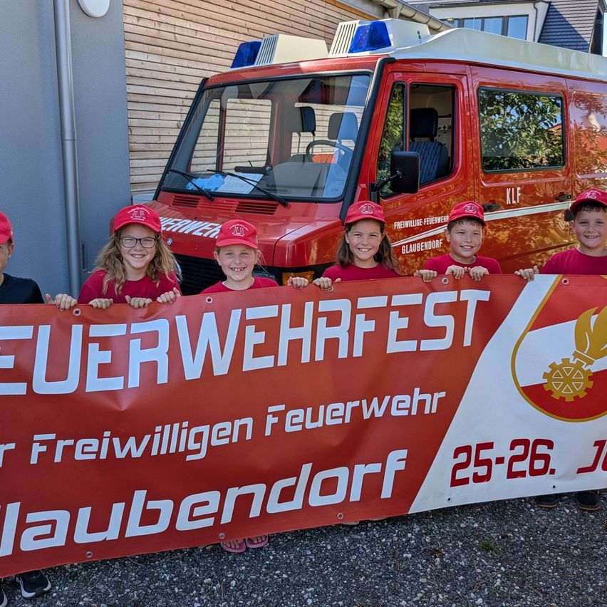 Eine Gruppe von Kindern in roten Mützen und T-Shirts steht hinter einem Banner, das 'Feuerwehrfest' vor einem roten Feuerwehrwagen besagt. Das Banner wird von den Kindern hochgehalten, und sie lächeln für die Kamera.