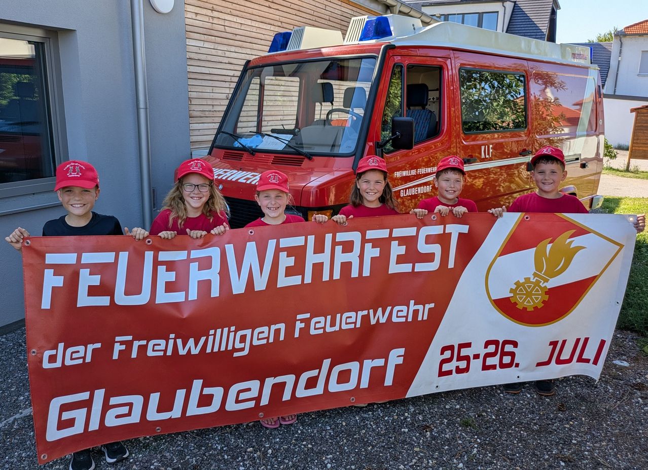 Eine Gruppe von Kindern in roten Mützen und T-Shirts steht hinter einem Banner, das 'Feuerwehrfest' vor einem roten Feuerwehrwagen besagt. Das Banner wird von den Kindern hochgehalten, und sie lächeln für die Kamera.