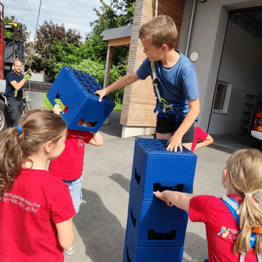 Eine Gruppe von Kindern in roten T-Shirts stapelt blaue Kisten, während ein Mann in einem blauen Shirt und Gurt zuschaut.