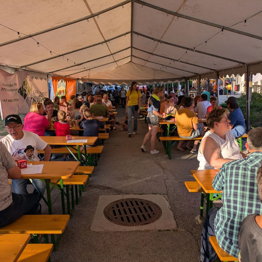 Eine Gruppe von Menschen sitzt unter einem Zelt an Tischen, einige essen, während andere herumstehen. Es hängen Banner am Zelt.