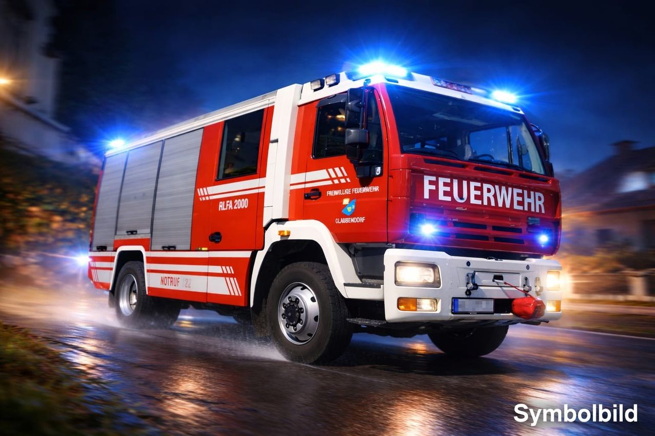 Ein roter Feuerwehrwagen mit dem Wort 'Feuerwehr' auf der Front fährt bei Nacht auf einer nassen Straße mit eingeschalteten Scheinwerfern und Blaulicht.