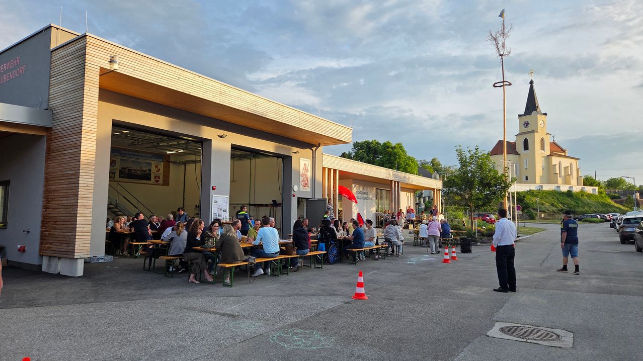 Sitzbereich im Freien mit mehreren Tischen und Bänken. Leute sitzen und stehen um die Tische. Das Gebäude hat eine rote Fahne. Bäume und ein rotes Auto sind im Hintergrund.