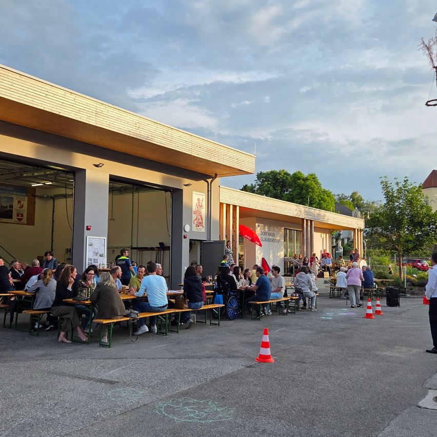 Sitzbereich im Freien mit mehreren Tischen und Bänken. Leute sitzen und stehen um die Tische. Das Gebäude hat eine rote Fahne. Bäume und ein rotes Auto sind im Hintergrund.