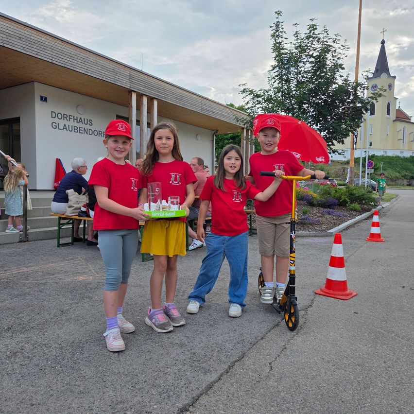 Vier Kinder in gleichen roten T-Shirts posieren für ein Foto vor einem Gebäude, eines hält eine Schale und ein anderes fährt einen Roller.