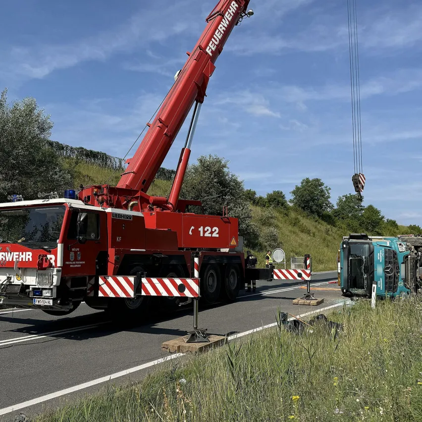 Ein Liebherr-Kran steht an der Straße und hebt ein beschädigtes grünes Fahrzeug an. Er ist neben der Straße mit einer Barriere geparkt. Im Hintergrund befinden sich Bäume und Gras.
