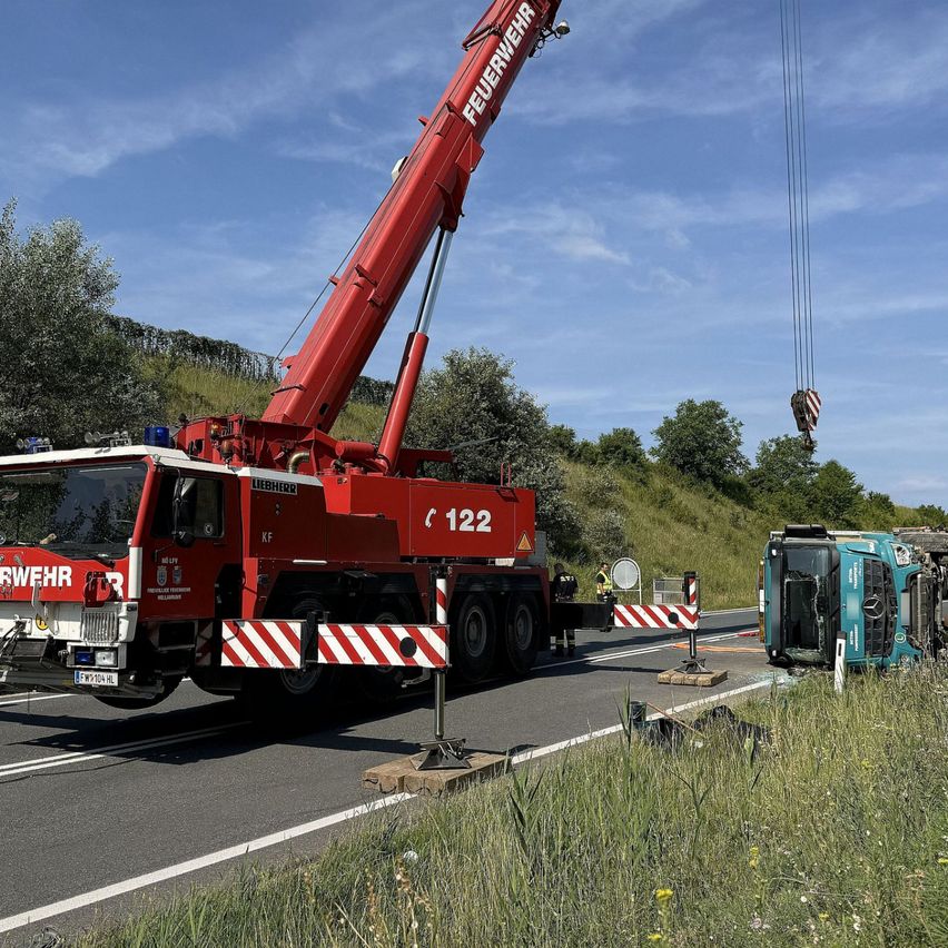 Ein Liebherr-Kran steht an der Straße und hebt ein beschädigtes grünes Fahrzeug an. Er ist neben der Straße mit einer Barriere geparkt. Im Hintergrund befinden sich Bäume und Gras.