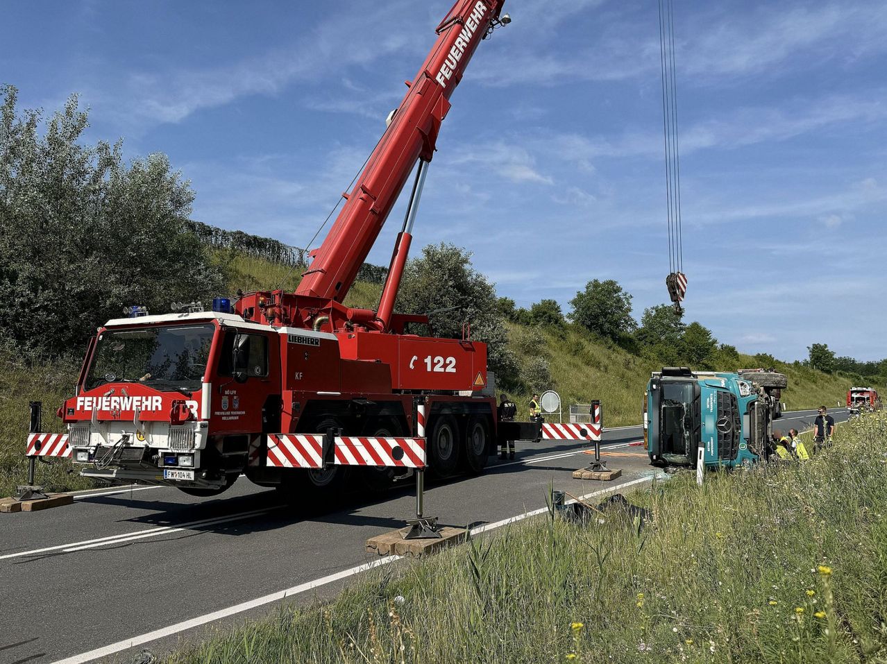 Ein Liebherr-Kran steht an der Straße und hebt ein beschädigtes grünes Fahrzeug an. Er ist neben der Straße mit einer Barriere geparkt. Im Hintergrund befinden sich Bäume und Gras.