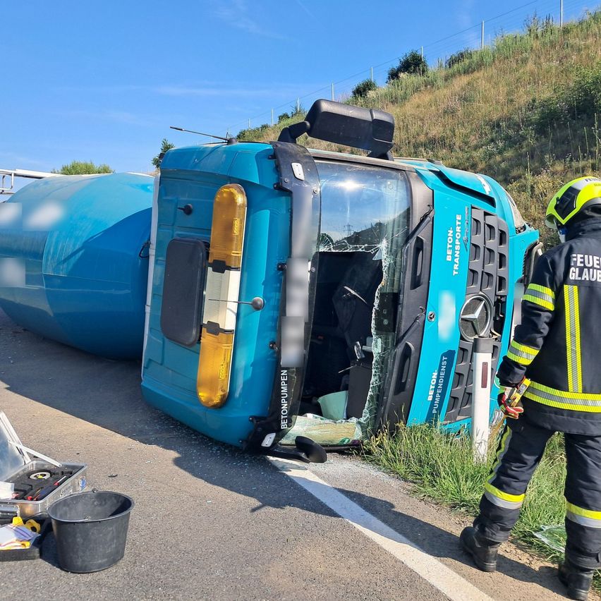 Ein blauer LKW mit beschädigter Front liegt auf der Straßenseite umgedreht, neben ihm steht ein Feuerwehrmann.