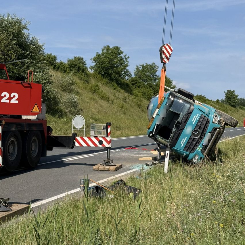 Ein blauer LKW liegt auf der Seite der Straße. Ein roter Kran hebt ihn hoch. Ein Straßenschild ist in der Nähe.
