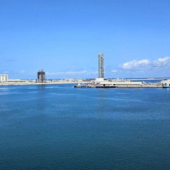 Ein Hafen mit Booten und Gebäuden. Ein großer Turm sticht in der Ferne hervor. Das Wasser ist ruhig.
