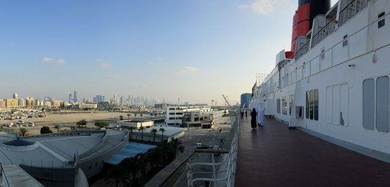 Ein Panoramablick vom Schiffsdeck zeigt eine moderne Stadtlandschaft mit Wolkenkratzern unter einem klaren Himmel, einem entfernten Hafen mit Schiffen und einem Gehweg mit zwei Personen.