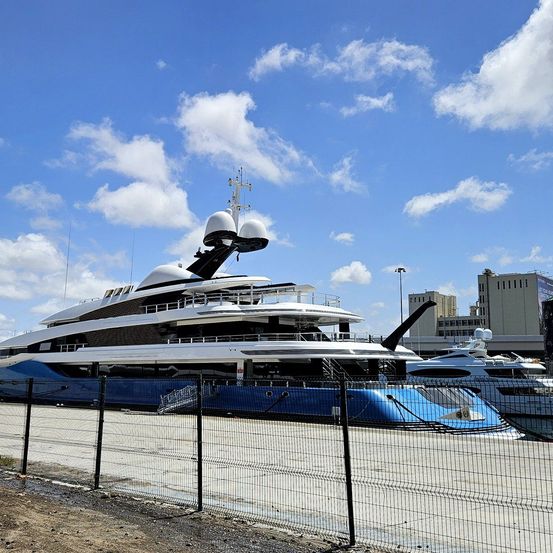 Ein blau-weißer Yacht liegt unter blauem Himmel mit Wolken, eingezäunt von einem Zaun. In der Ferne sind Gebäude und Straßenlaternen sichtbar.