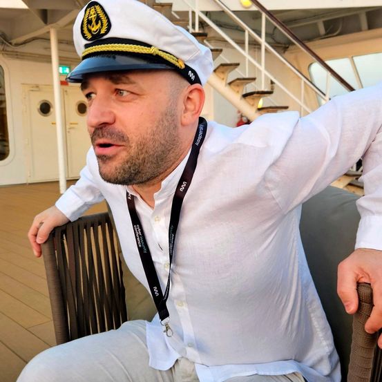 Ein Mann in einem weißen Uniform und einer dunkelblauen Mütze mit goldenem Ankersymbol sitzt auf einem Stuhl auf dem Deck eines Kreuzfahrtschiffs.