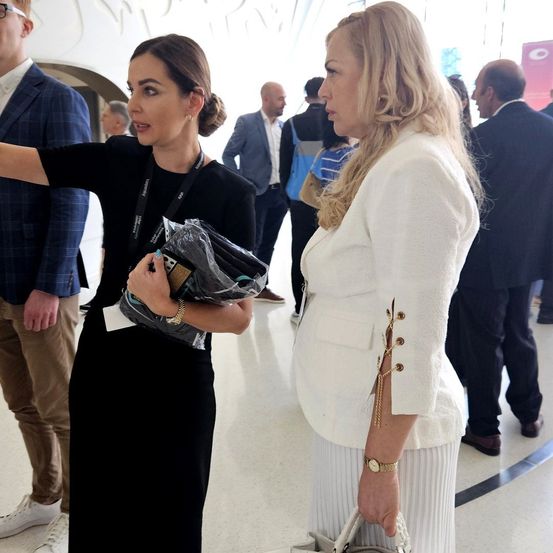 Zwei Frauen unterhalten sich in einem geschäftigen Flur, umgeben von Menschen. Eine Frau hält eine Tasche und trägt ein schwarzes Kleid; die andere, eine weiße Jacke.