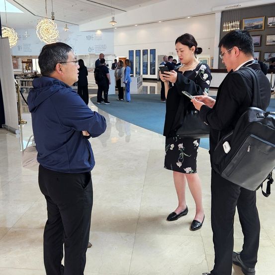 Drei Menschen stehen in einer Lobby, eine Frau schaut auf ihr Handy. Sie sind von Menschen umgeben, die ebenfalls ihre Telefone benutzen.