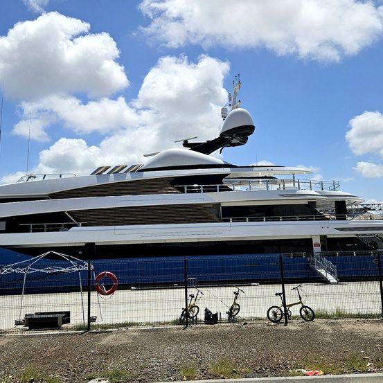 Ein großer blau-weißer Yacht ist in einem Hafen verankert. Fahrräder sind vor dem Yacht geparkt, und ein Rettungsring ist in der Nähe platziert.