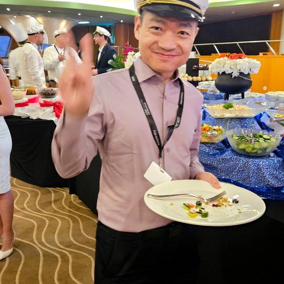 Ein Mann in Uniform hält einen Teller mit Essen und einer Gabel und macht ein Friedenszeichen. Dahinter steht eine Buffet-Tabelle mit verschiedenen Speisen und Leuten.
