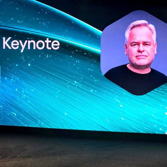 Ein Mann erscheint auf einem großen Bildschirm mit dem Wort 'Keynote'. Der Mann sieht ernst aus und der Bildschirm hat ein blaues, welliges Design.