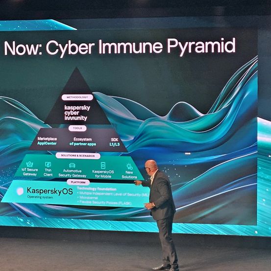 Ein Mann steht auf der Bühne und spricht zu einem Publikum, mit einer großen Leinwand hinter ihm, die die Cyber Immune Pyramid von Kaspersky zeigt.