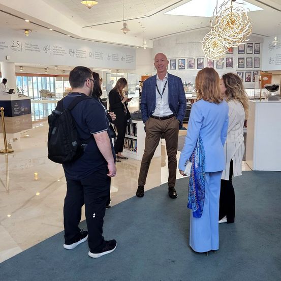 Eine Gruppe von Menschen befindet sich in einer Lobby. Ein Mann in einem blauen Anzug spricht mit einer Gruppe von Menschen. Eine Frau in einem blauen Mantel hält eine Handtasche.