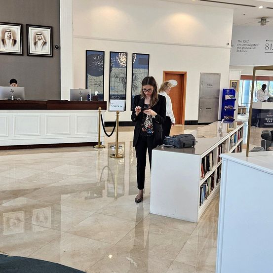 Eine Frau schaut auf ihr Handy in einer leeren Lobby. Es gibt einen Schreibtisch mit Laptop, Bücher auf einem Regal und gerahmte Bilder an der Wand.