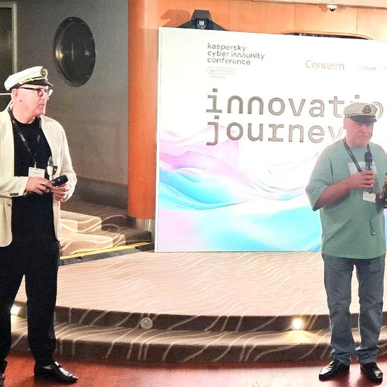 Zwei Männer stehen auf der Bühne, einer mit einem Mikrofon und einem Getränk, der andere mit einem Telefon und einem Glas. Hinter ihnen steht ein großes Banner mit der Aufschrift 'Kaspersky Cyber Immunity Conference'.