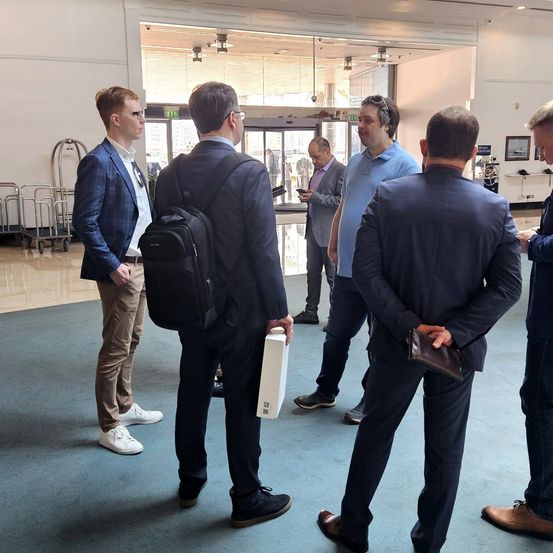 Fünf Männer in Anzügen stehen in einer Lobby. Einer hält eine weiße Box. Ein Mann in einem beigen Anzug hält einen Rucksack.