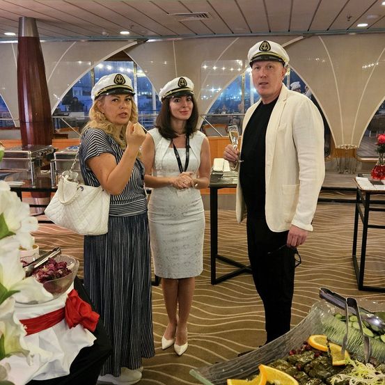 Drei Personen in einem Raum mit einem Buffettisch. Zwei Frauen in Hüten und ein Mann in Anzug. Frau auf der linken Seite hält eine weiße Tasche.